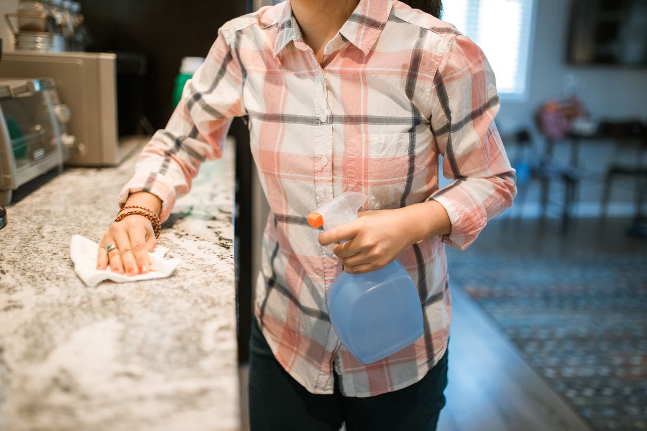 gallery-03 A woman wearing a plaid shirt cleans a kitchen counter using a spray bottle and cloth.
