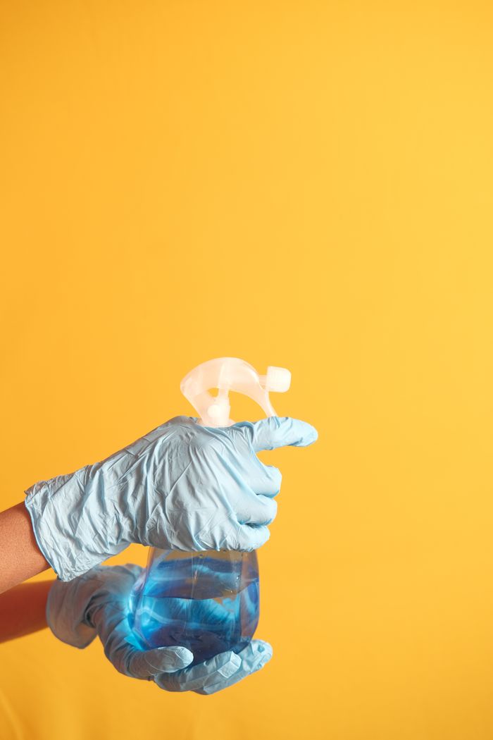 gallery-01 Hand in blue glove holding a cleaning spray bottle against a yellow background.