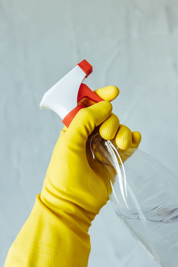 home-img A hand wearing a yellow rubber glove holds a red and white spray bottle against a neutral background.
