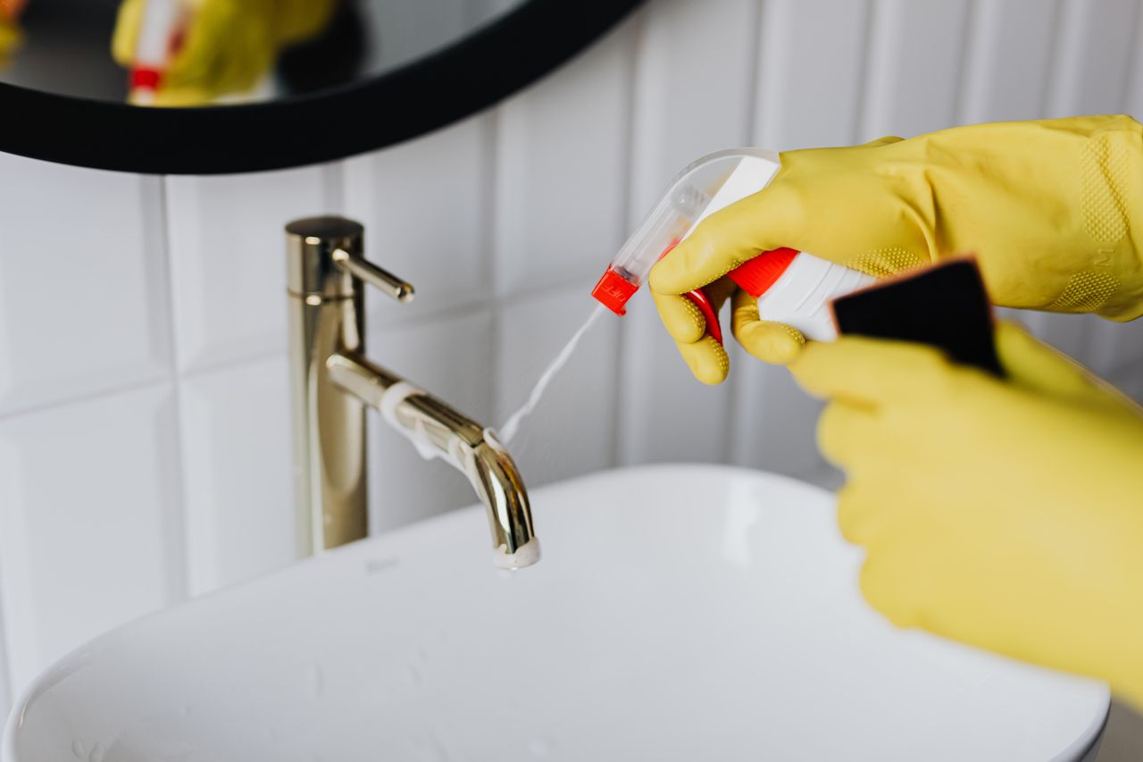 gallery-02 From above anonymous housekeeper in yellow rubber gloves spraying cleaner to chrome water faucet in bathroom